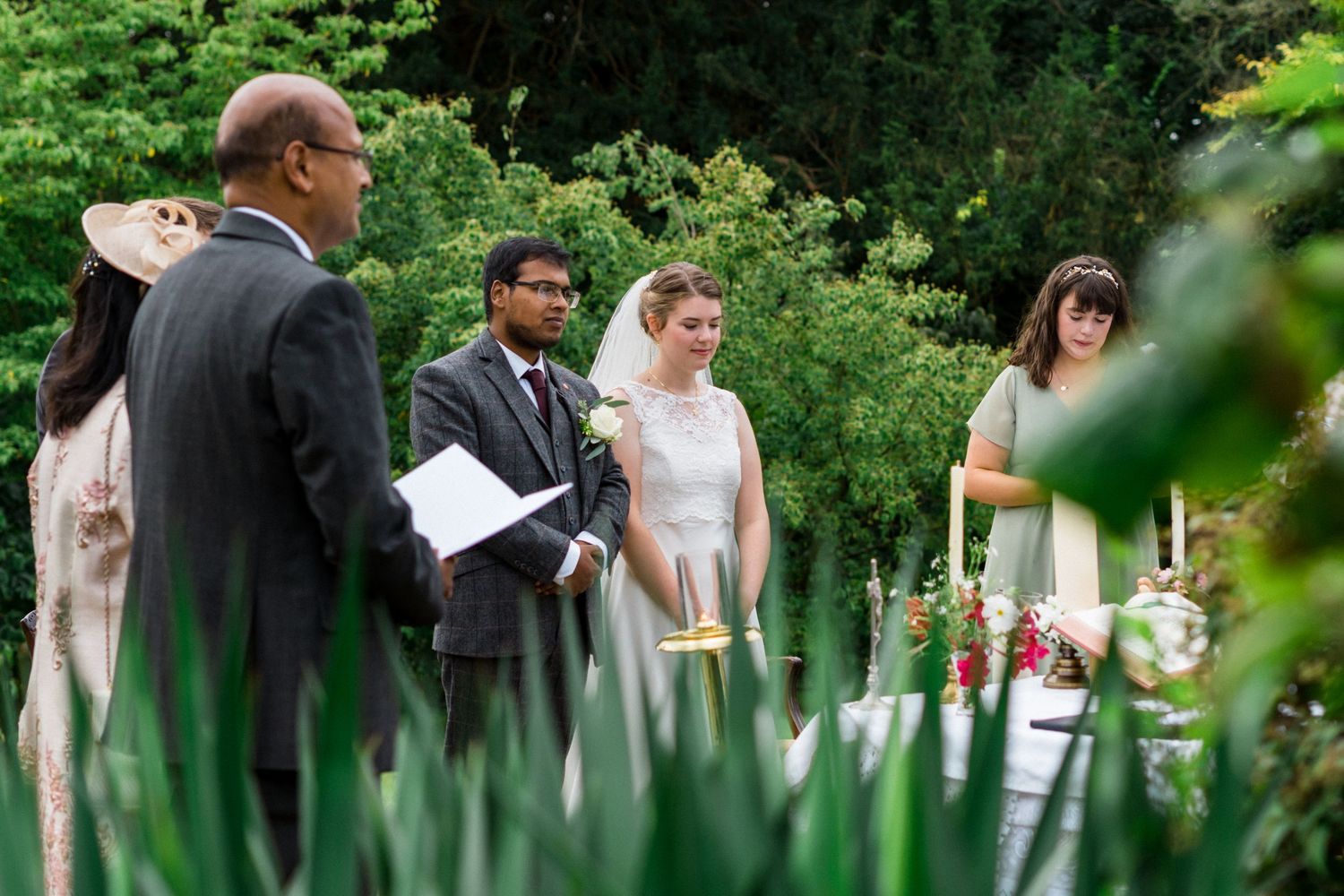 Outdoor garden wedding ceremony with bride and groom standing before officiant amid lush green foliage.