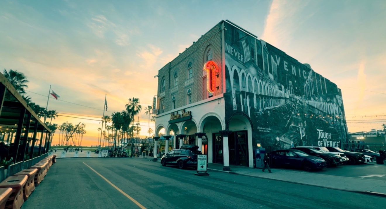 Vintage building with neon signs glows at sunset along a palm-lined street in a coastal town.