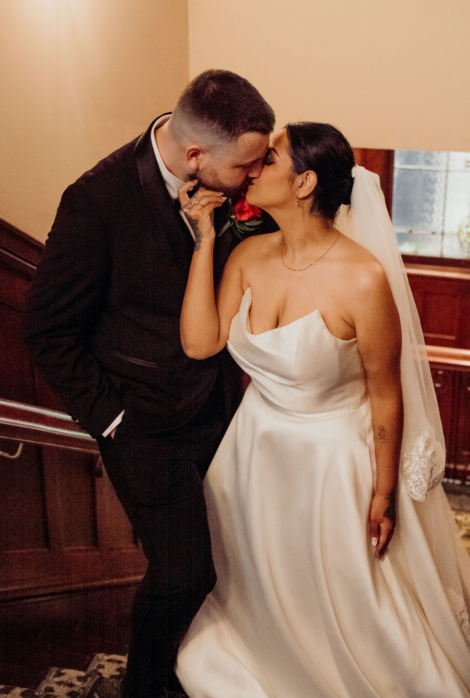 A bride and groom share a romantic kiss, she in a white gown with veil, he in a dark suit, holding a red rose.