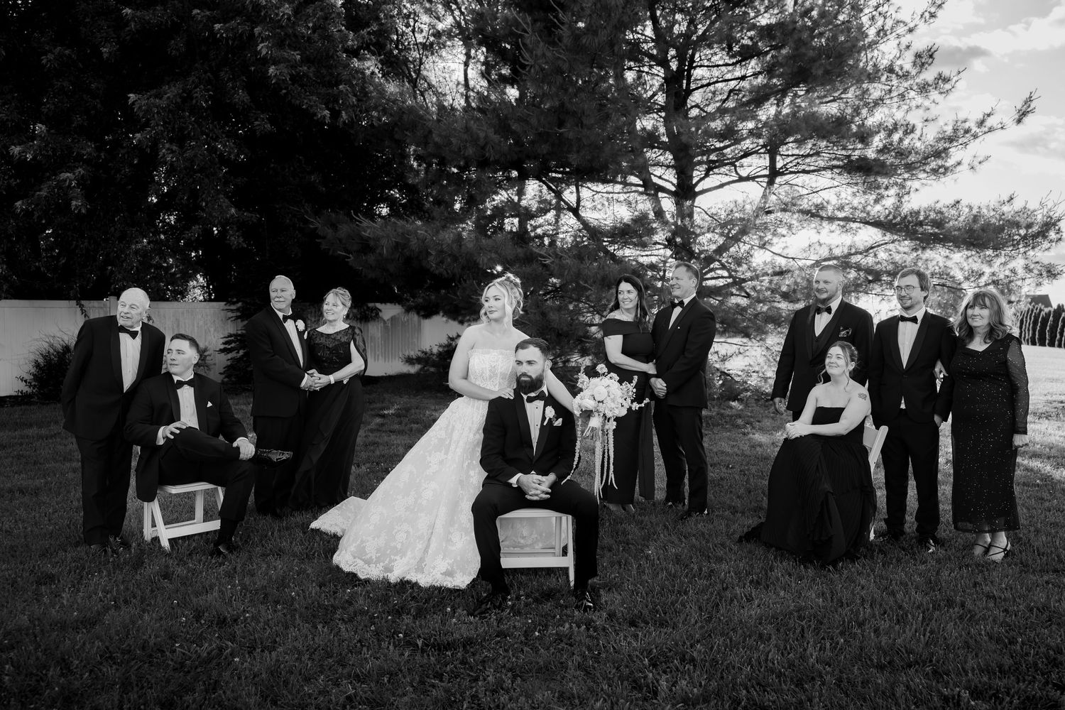 Black and white wedding photo of bride and groom with family group posed outdoors on lawn near trees.