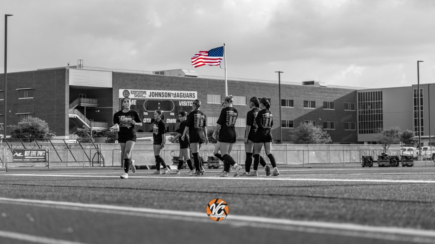 Soccer players in black uniforms walk on a field near an American flag outside Johnson school building.