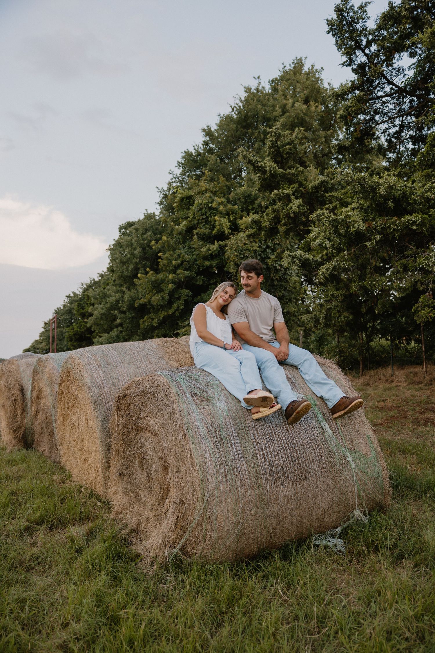 Couple sitting together on a large hay bale in a grassy field during golden hour.