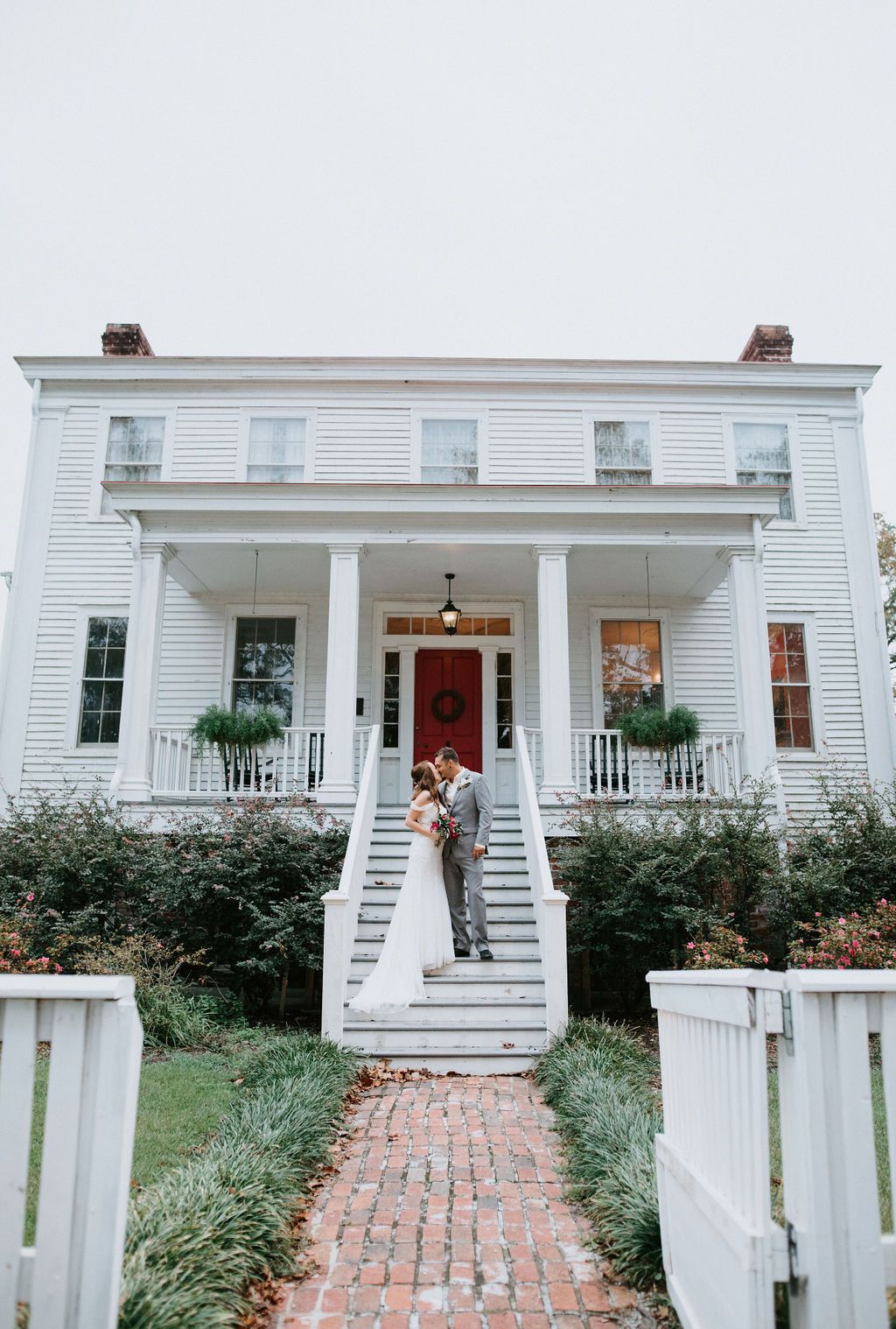 White colonial style house with brick walkway and pillared front porch decorated with hanging plants and red door.