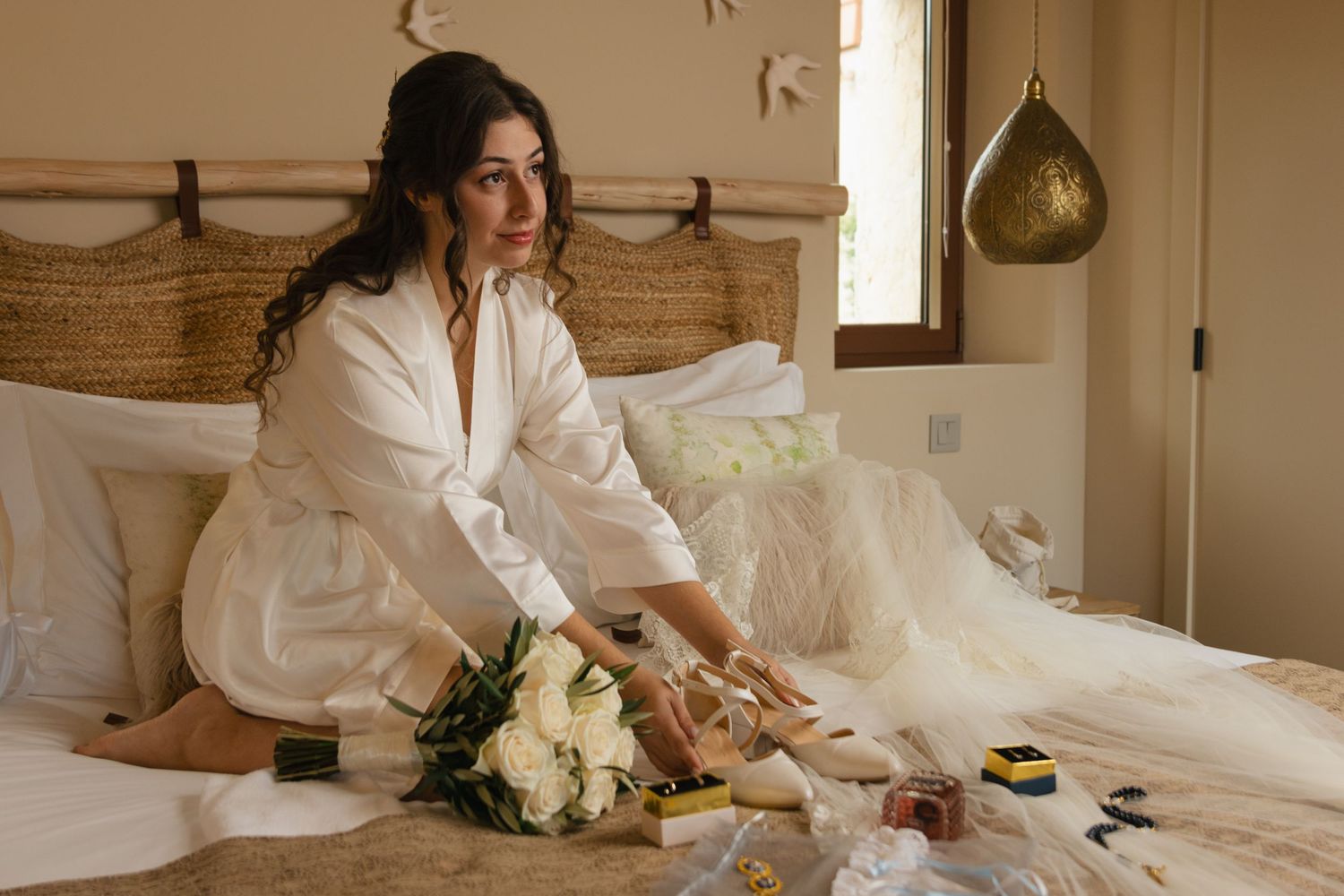 Someone in a white robe sits on a bed with a bouquet of white flowers and wedding dress nearby.