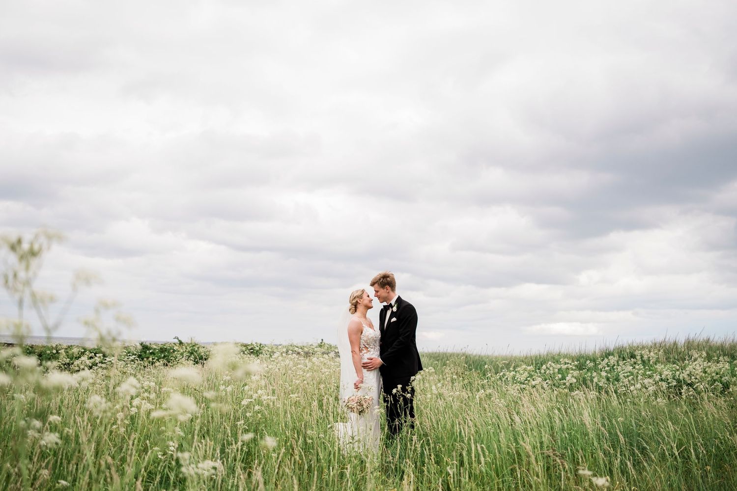 A bride and groom stand together in a lush wildflower meadow under a dramatic cloudy sky on their wedding day.