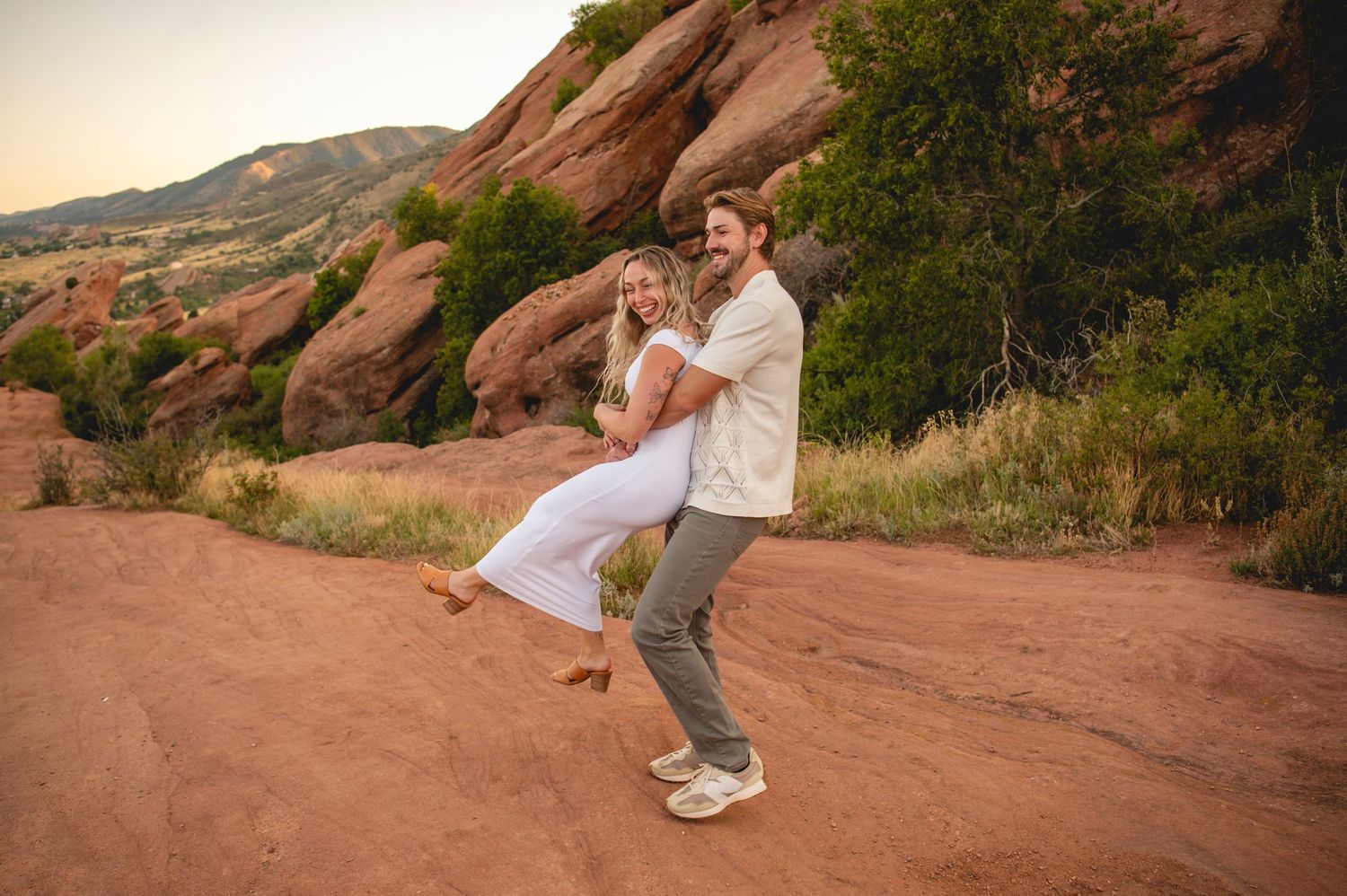 Couple dancing joyfully on red rock trail at Red Rocks Park, Colorado, during golden hour sunset.