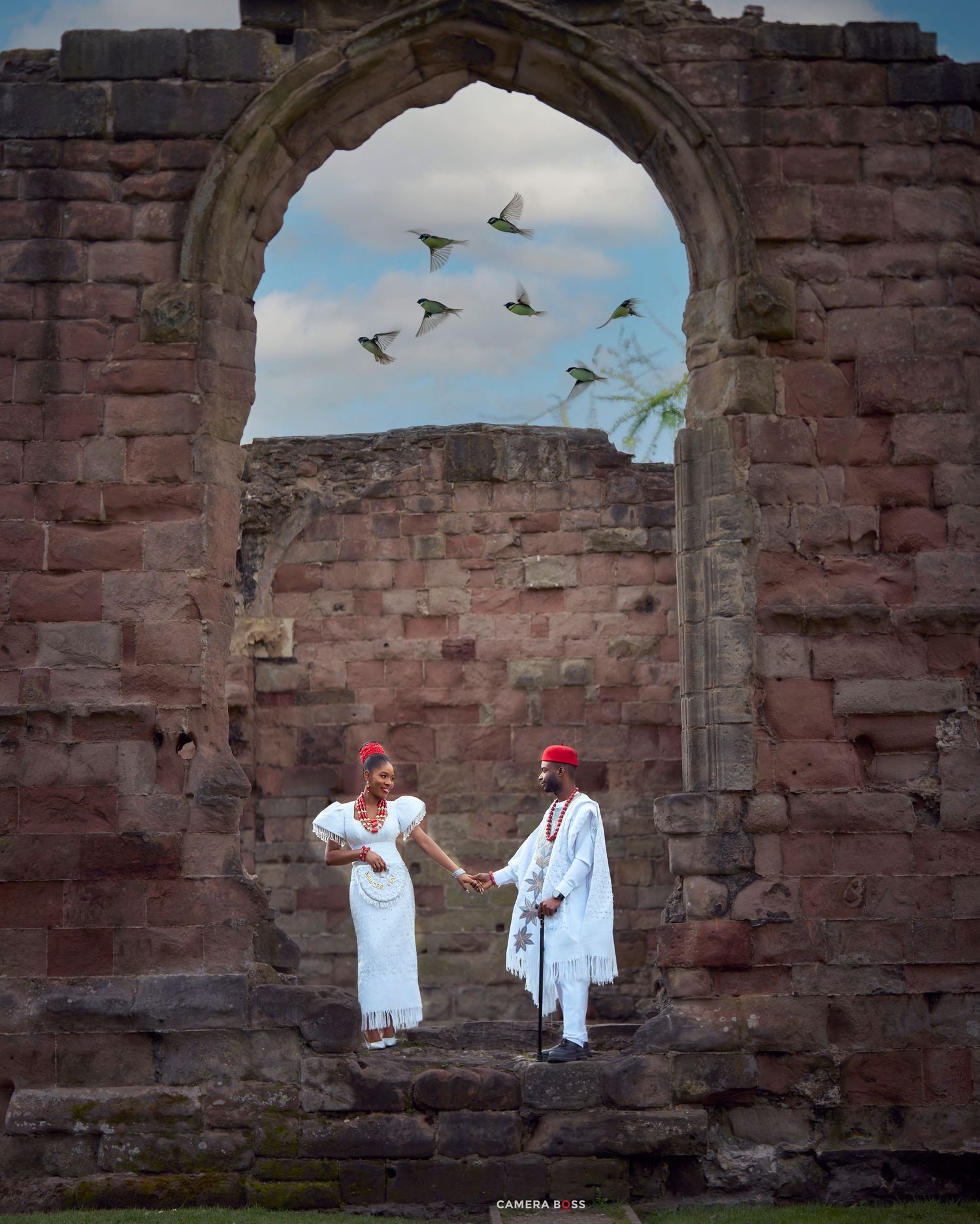 Couple in white traditional attire holding hands under a stone arch at ancient ruins with birds flying overhead.