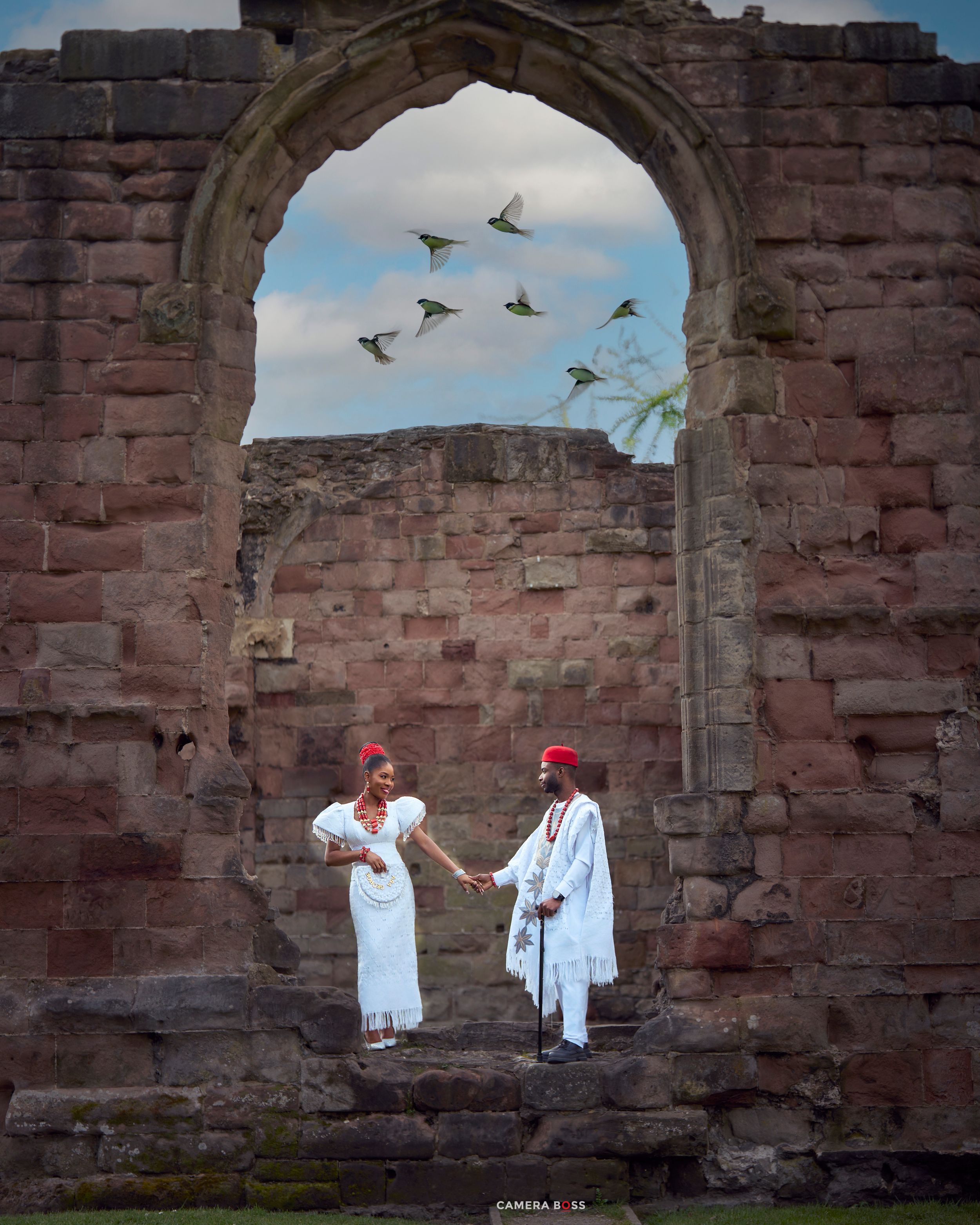 Couple in white traditional attire holding hands under a stone arch at ancient ruins with birds flying overhead.