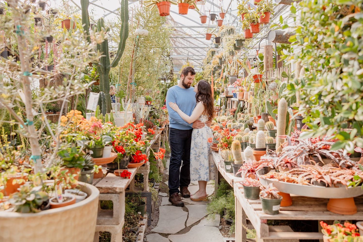Couple stands close together between rows of potted plants in a greenhouse, surrounded by colorful flowers.