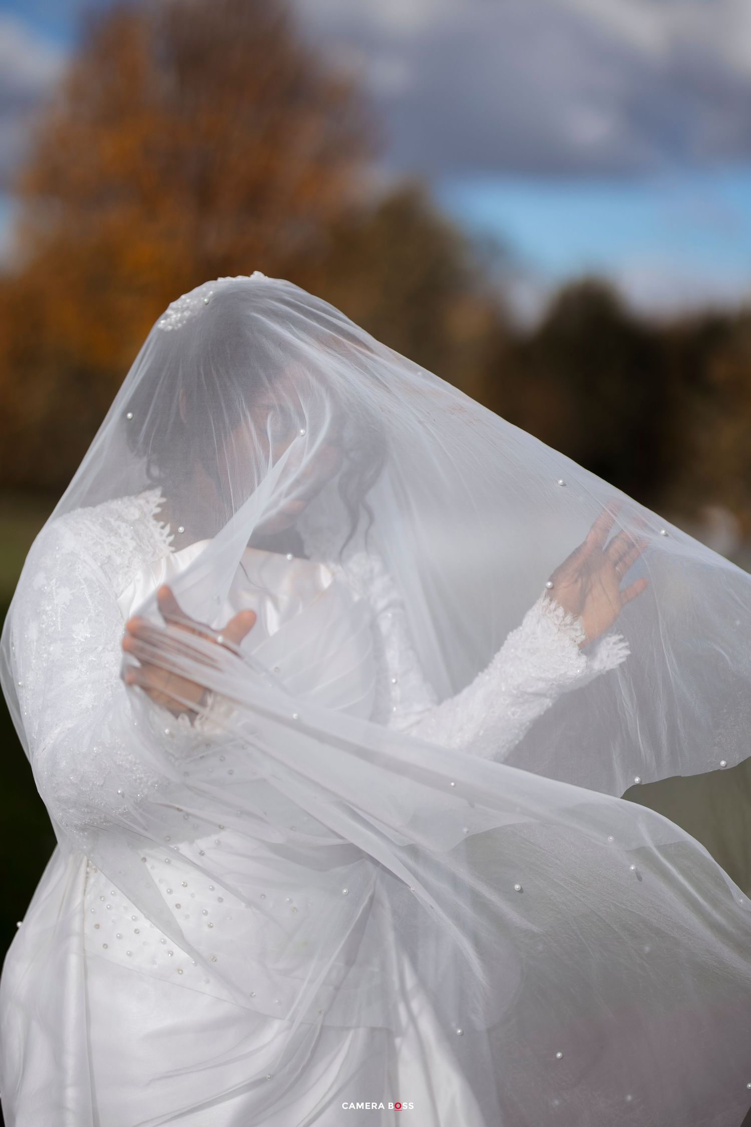 Bride in white lace gown with flowing veil blowing in wind, outdoor autumn wedding photo.