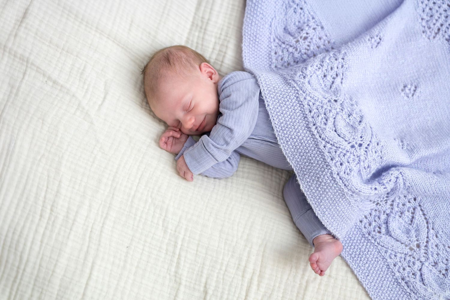 Sleeping newborn baby in a blue onesie wrapped in a light purple knit blanket on white bedding.