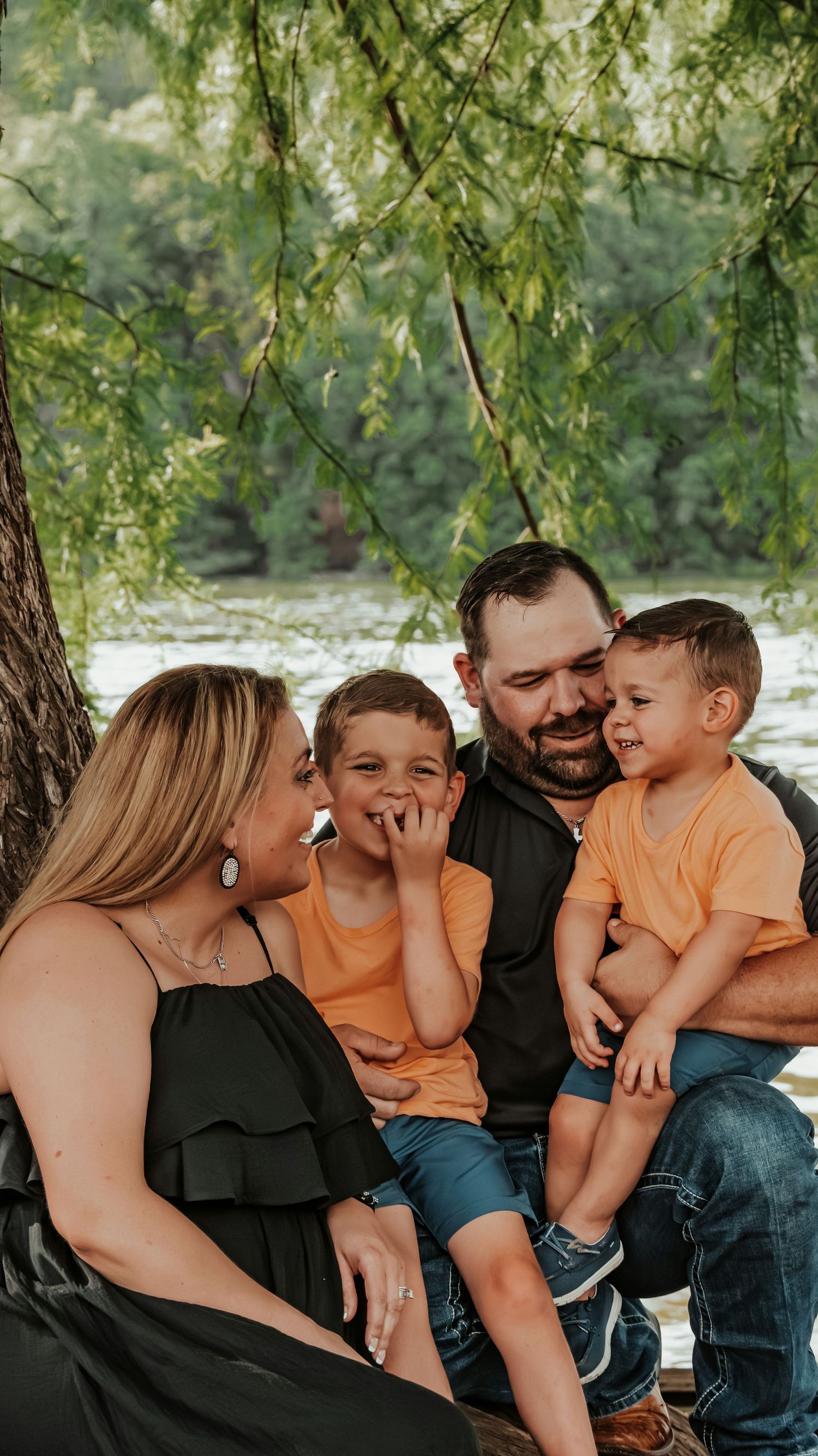 Family of four posing outdoors in a field, parents in black outfits with two young boys in matching orange shirts.