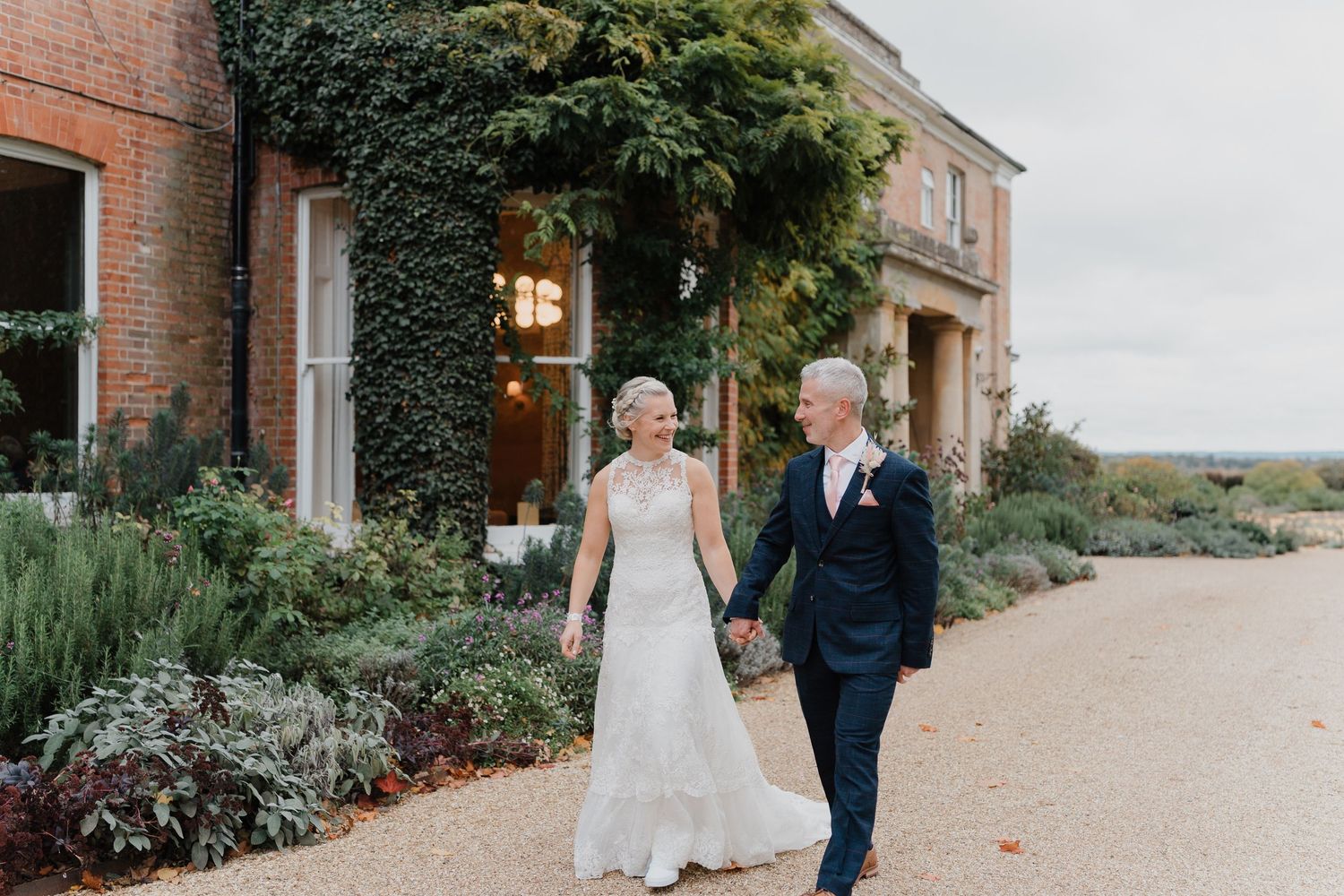 Bride and groom holding hands walking outside an ivy-covered brick wedding venue with lush gardens.