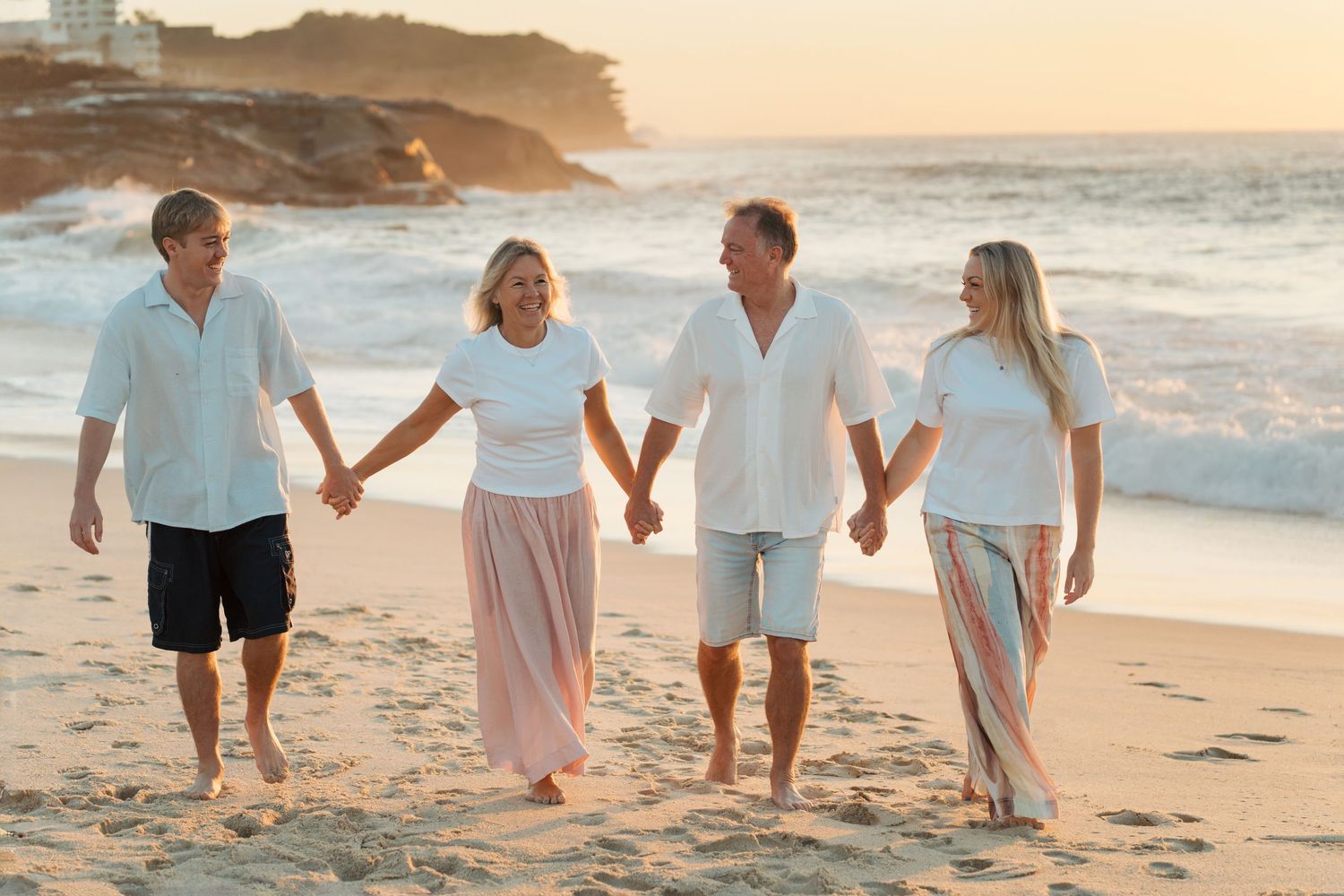 family of four holding hands walk along a sunny beach at sunrise wearing white tops and flowing skirts at Bronte beach family photoshoot
