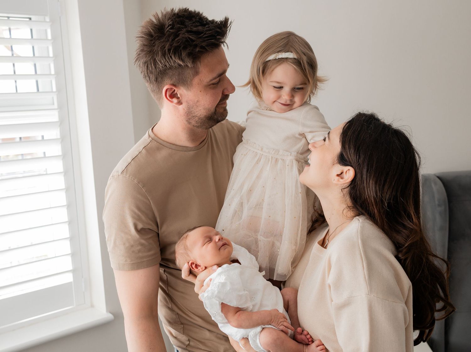 A happy family of four with a dad, mom, toddler girl, and newborn baby posing together near a bright window.