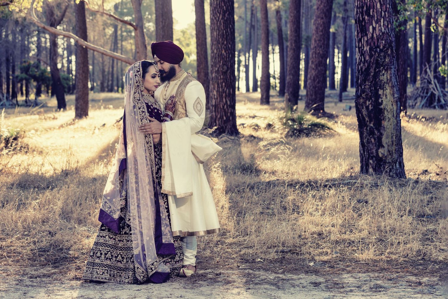 Couple in traditional South Asian wedding attire embracing in a sunlit pine forest.