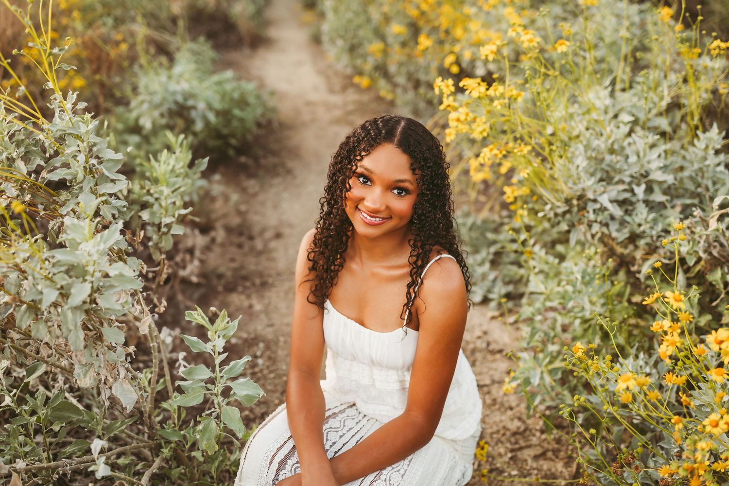Young woman in white dress smiling among yellow wildflowers on a sunny garden path.