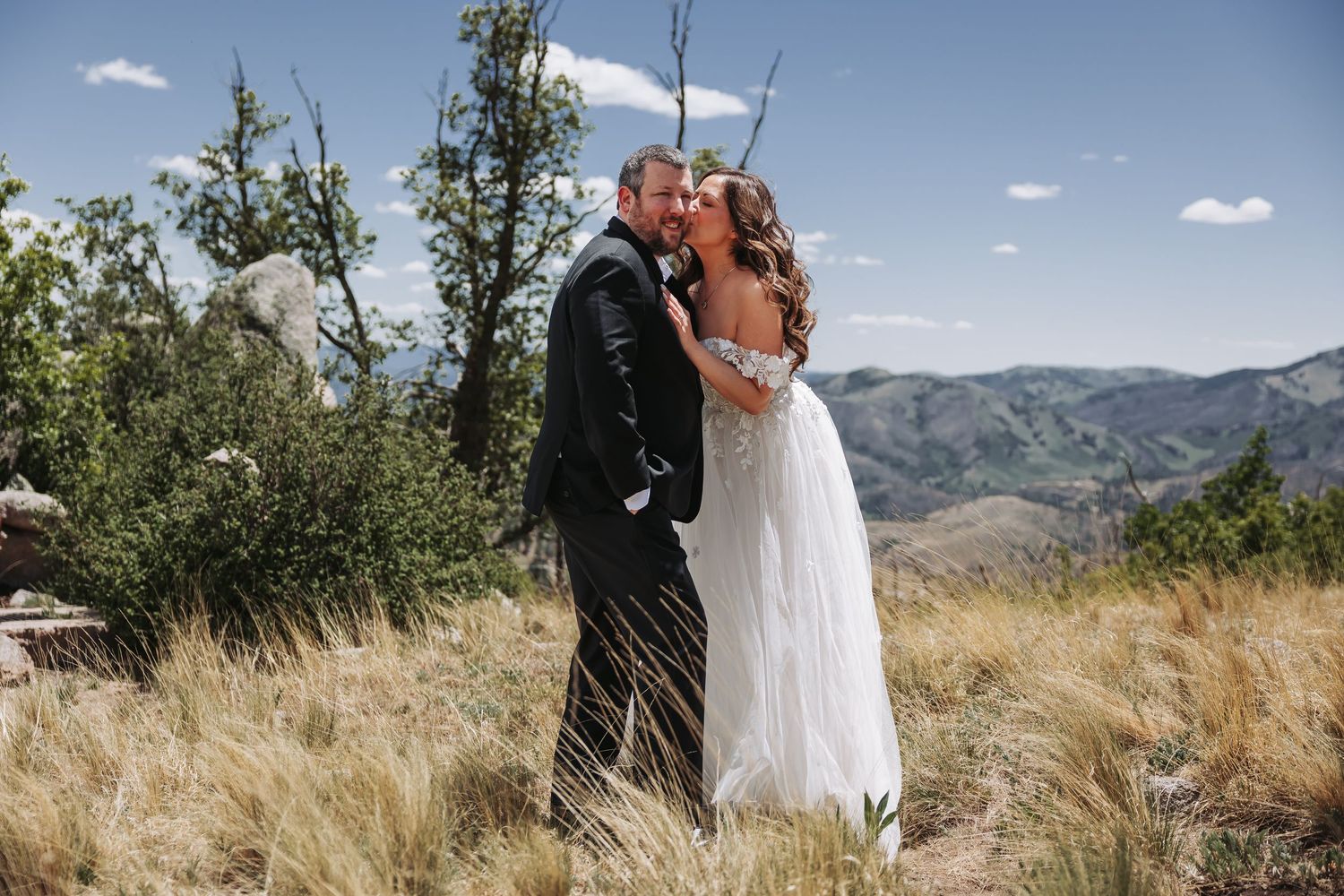 A couple shares a romantic kiss in wedding attire on a mountain overlook with scenic hills and blue sky.