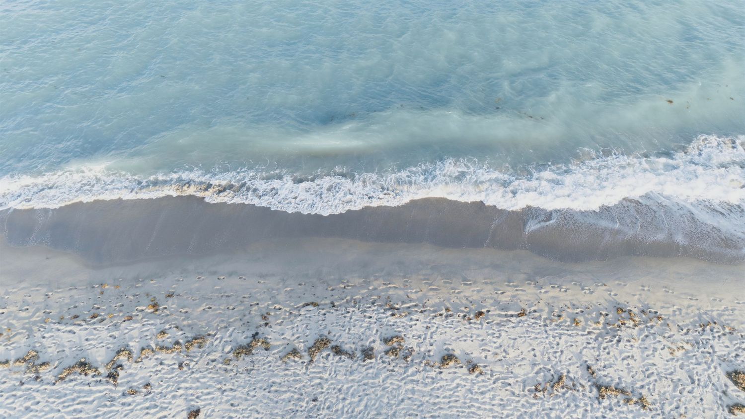 Aerial view of gentle ocean waves washing onto a rocky pebble beach with clear turquoise water.