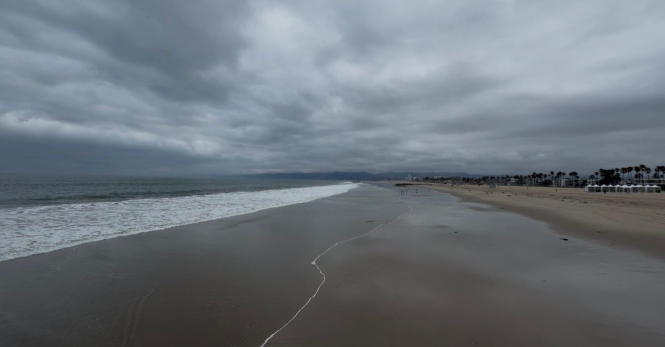Overcast stormy beach scene with dark clouds, crashing waves, and wet sand stretching along a coastal shoreline.