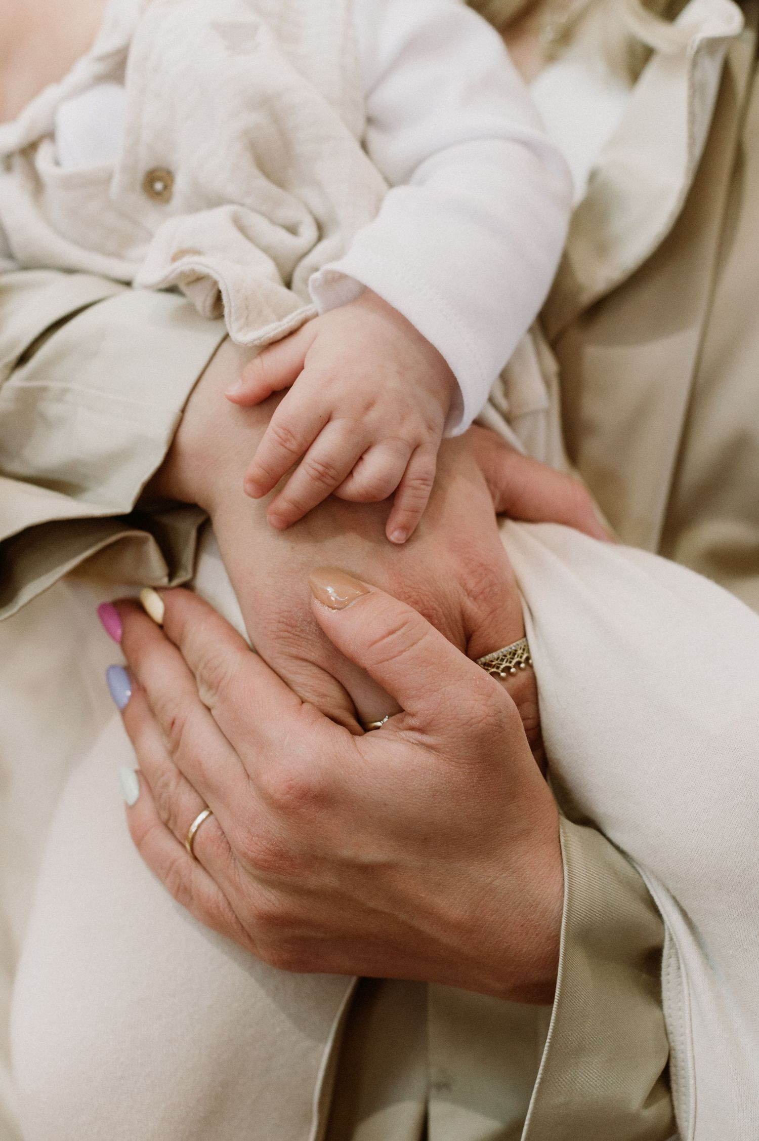 Close-up of a parent cradling a newborn baby's tiny hands, wearing rings, dressed in neutral tones.