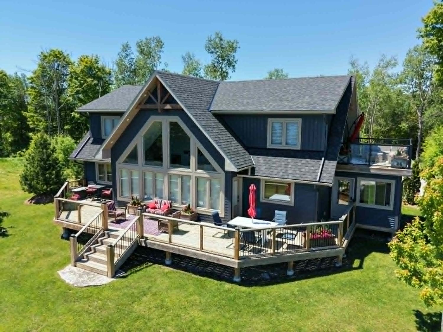 Aerial view of a large blue cottage with a spacious wooden deck surrounded by lush green trees on a sunny day.