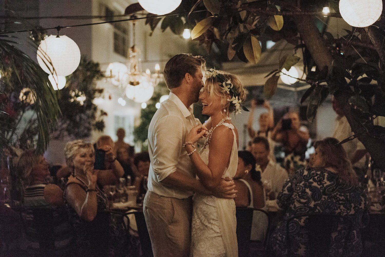 Couple sharing first dance in warmly lit tropical restaurant venue surrounded by guests and hanging globe lights.