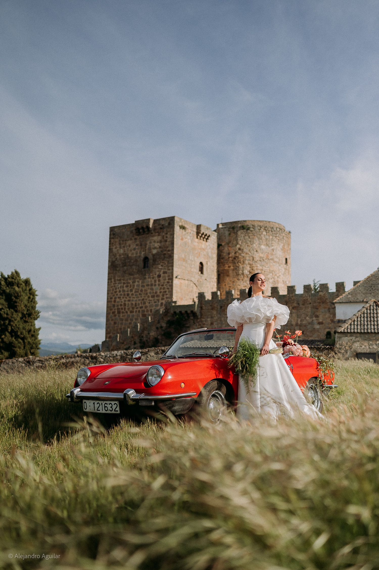 Bride in a white off-shoulder gown posing beside a red vintage convertible car near a medieval castle.