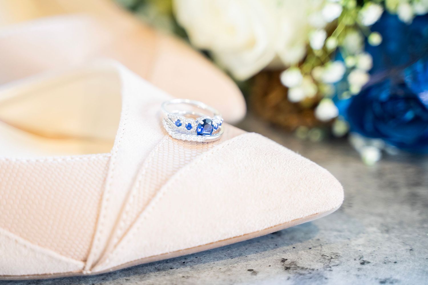 Elegant blush wedding shoe with a sapphire and diamond ring resting on top.