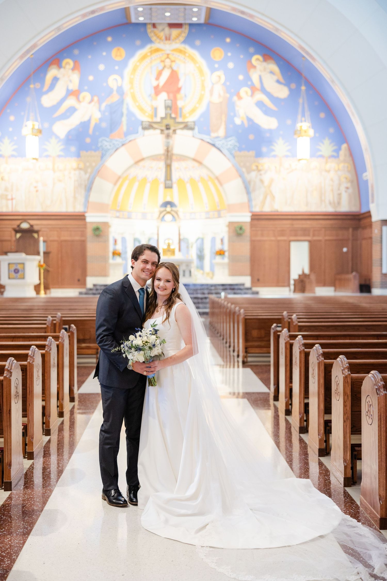 Wedding couple poses in elegant church with beautiful painted mural and wooden pews.
