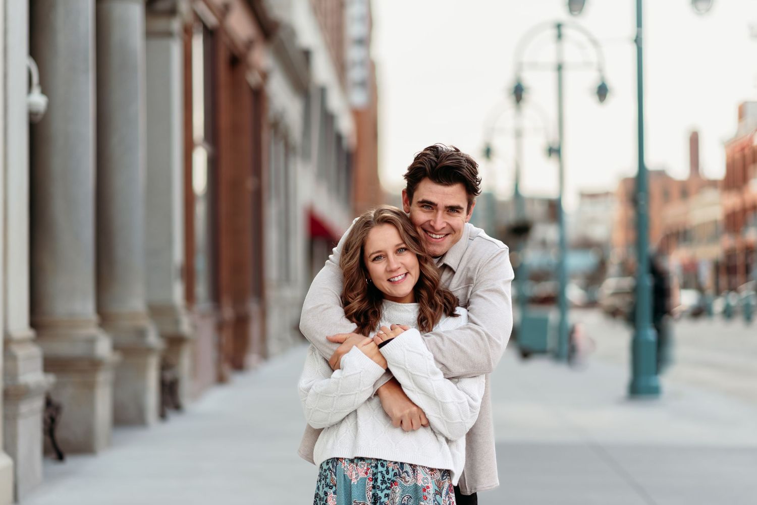 A couple embraces on a city street with classic architecture and lamp posts in the background.