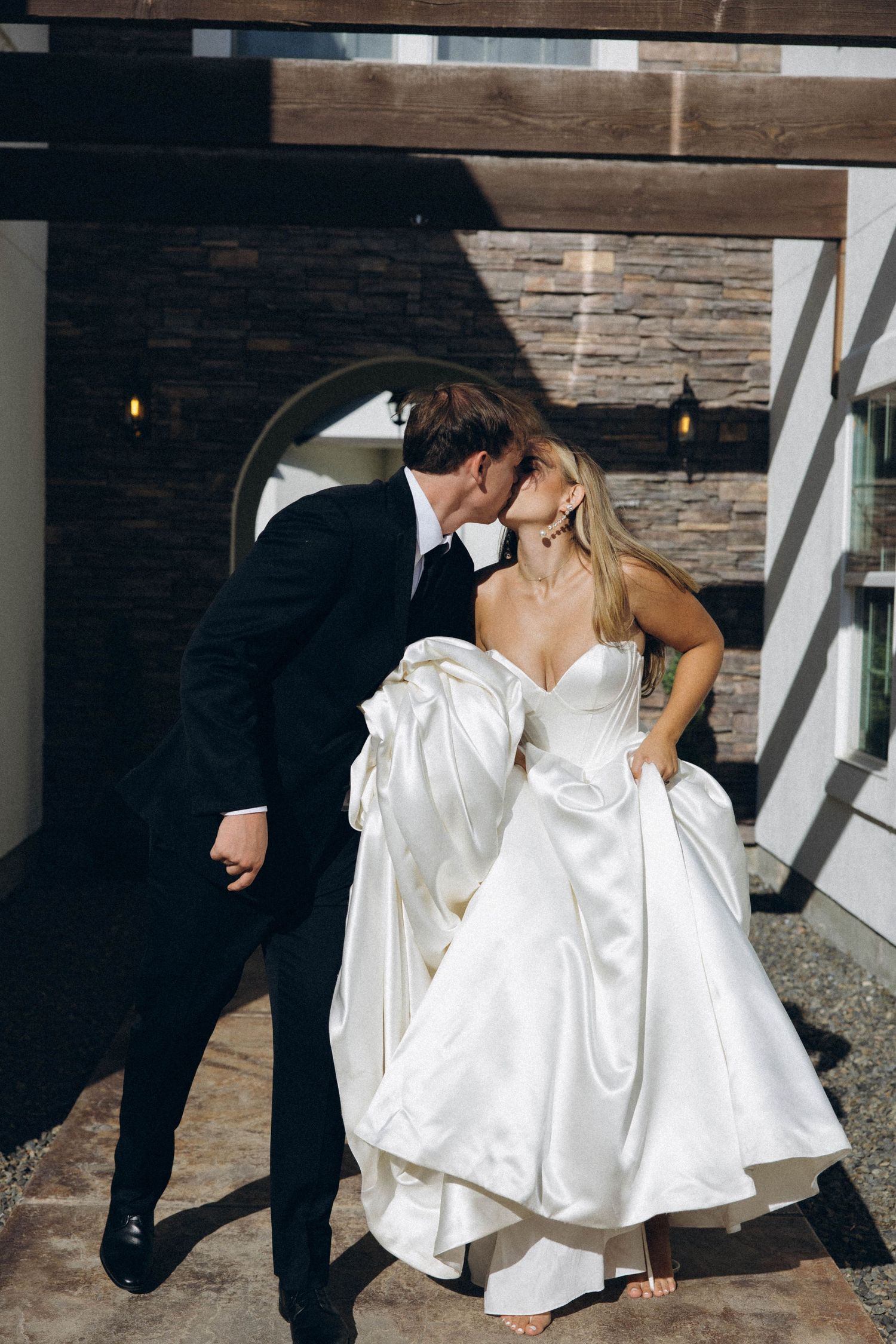 Elegant couple shares intimate moment in white gown and black suit against rustic stone wall.