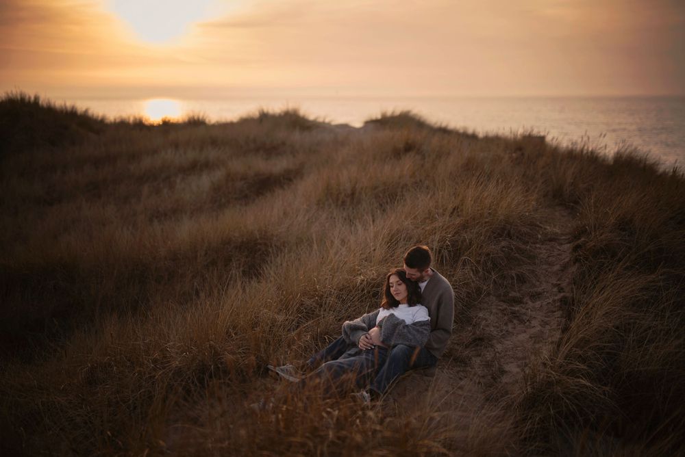 A couple sits on a grassy hillside at sunset overlooking the ocean in a romantic outdoor setting.