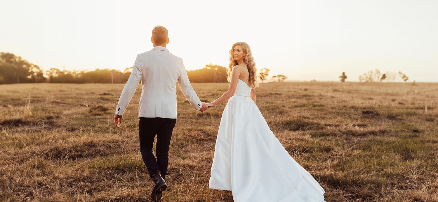 Romantic sunset photograph of newlyweds walking hand in hand in a golden field.