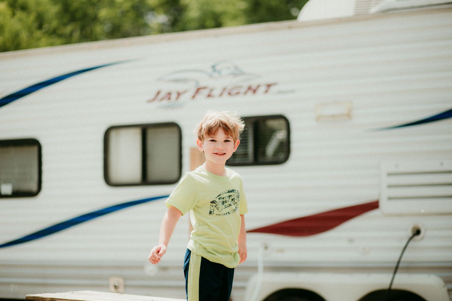 A young child in a green t-shirt stands in front of a white and blue Jayfeather travel trailer at a campground.