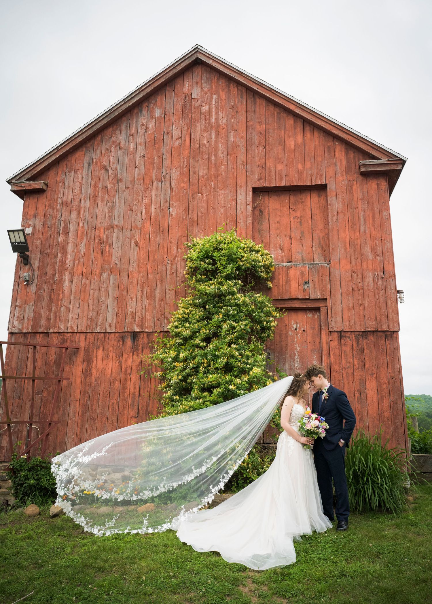 Romantic wedding portrait series showing couple in formal attire posing against rustic red barn with veil flowing in wind.
