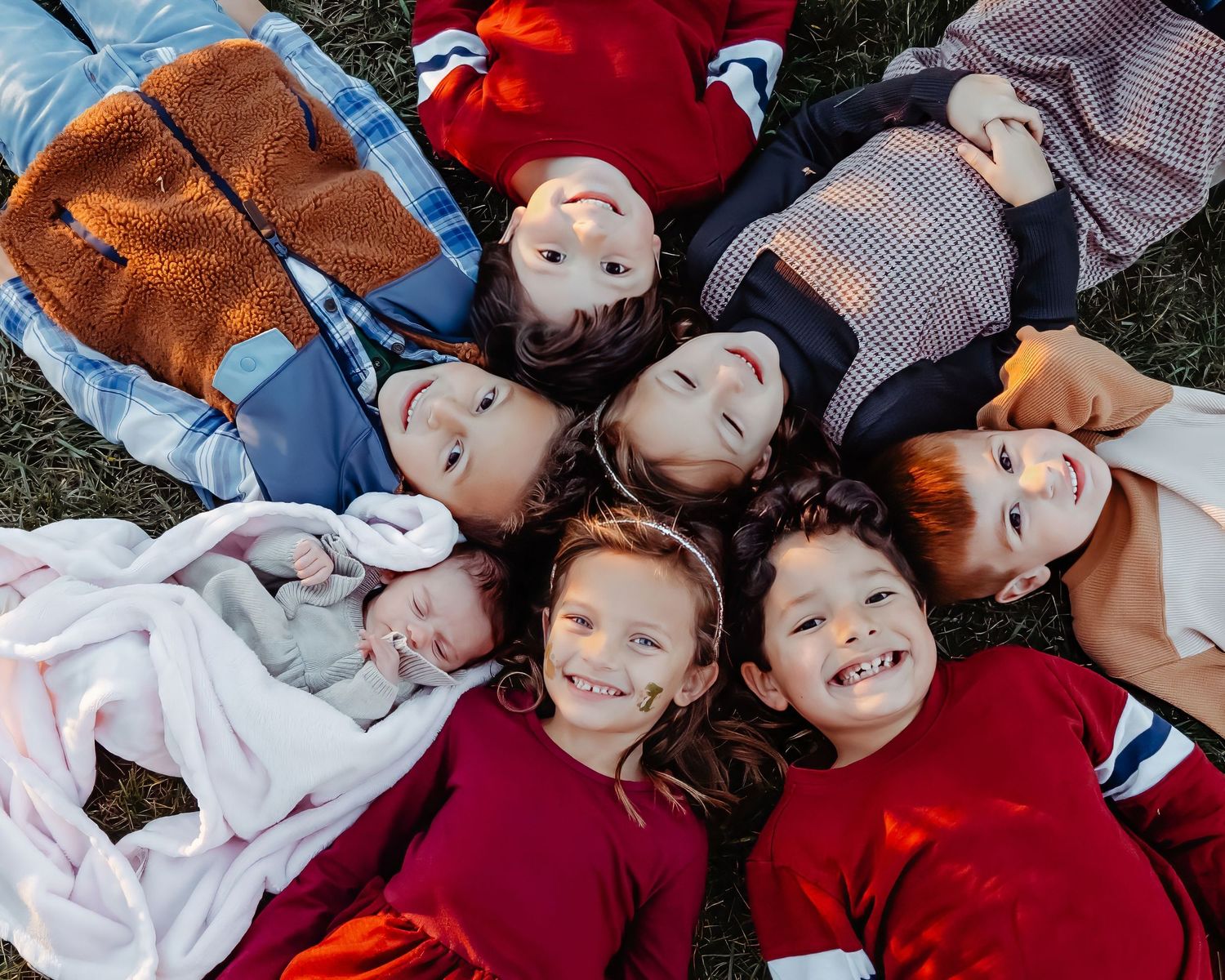 A group of children lying in a circle on grass wearing casual clothing smiling up at the camera.