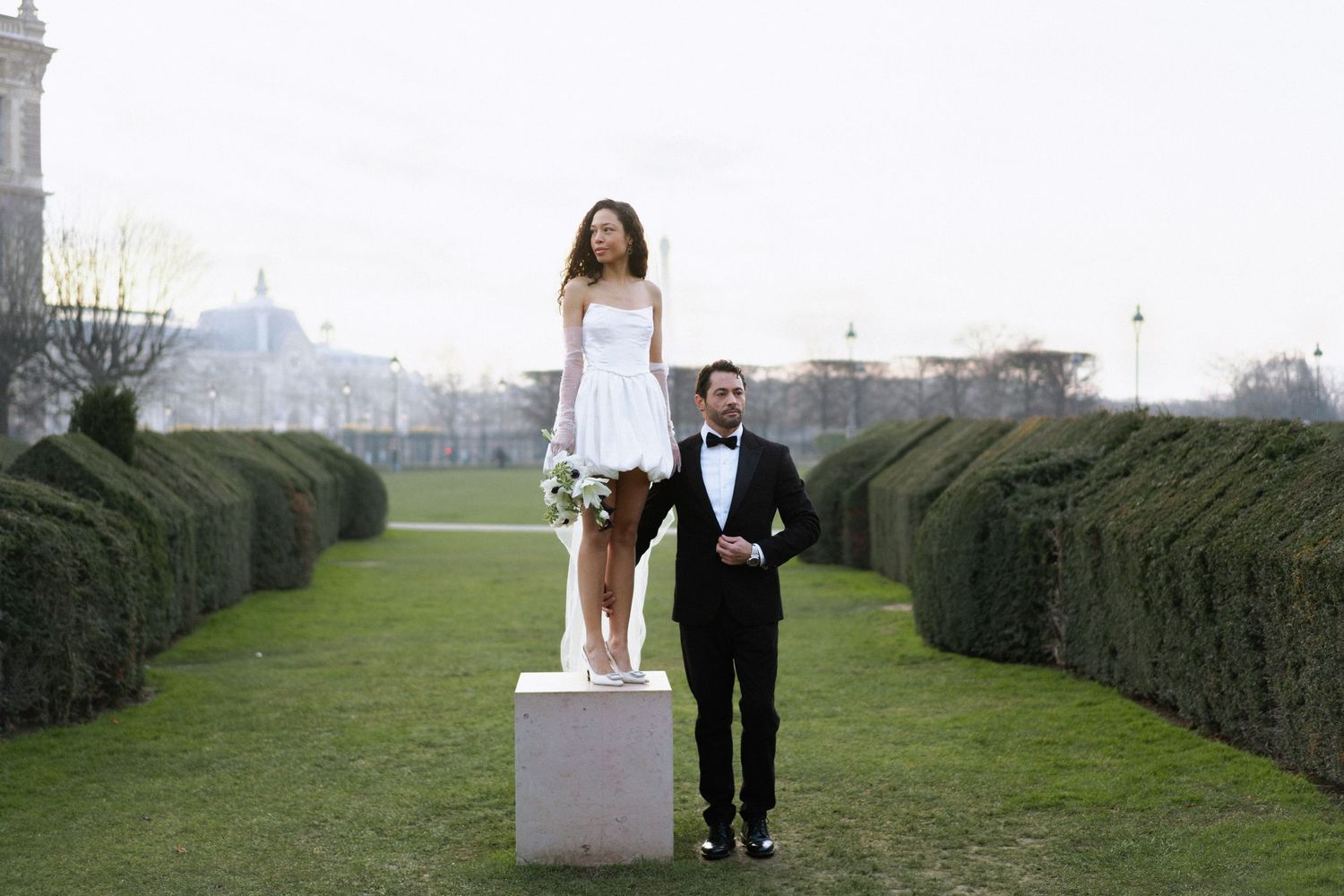 A couple poses creatively in a formal garden with manicured hedges, with one standing on a white pedestal.