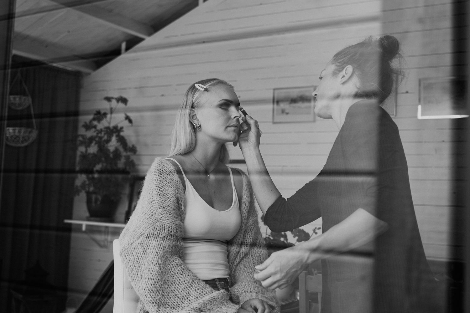 Makeup-artist sminkar kvinna i ljus tröja, sett genom fönster i svartvitt. Fotograf Gabriel Alenius, Falun