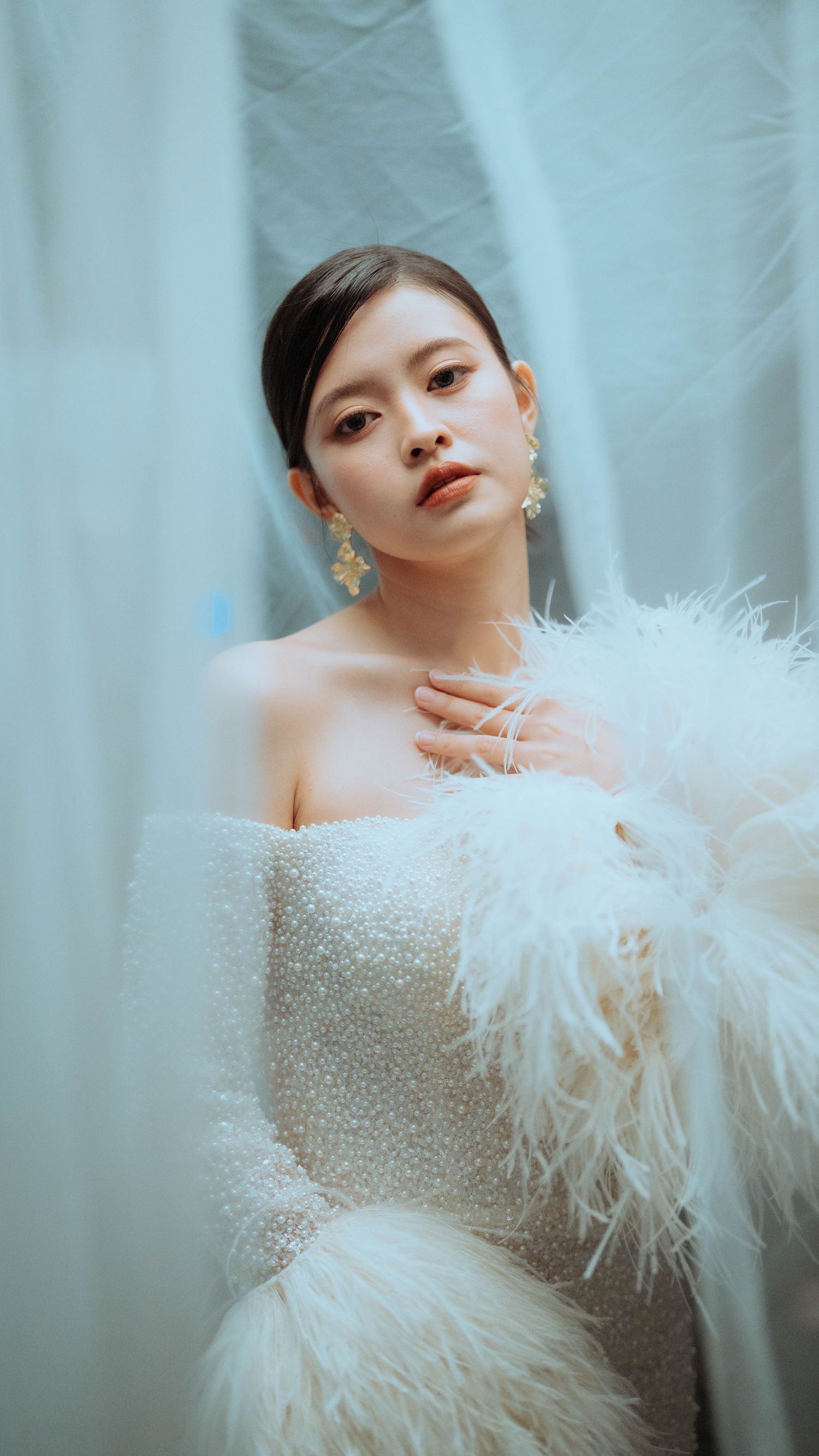 A glamorous bridal portrait featuring an elegant white beaded gown with feather details against a soft blue backdrop.