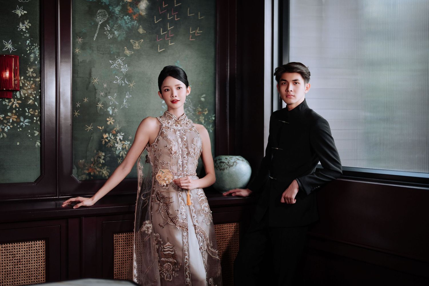 Elegant couple in traditional Asian formal wear pose in a room with ornate wallpaper and vintage decor.