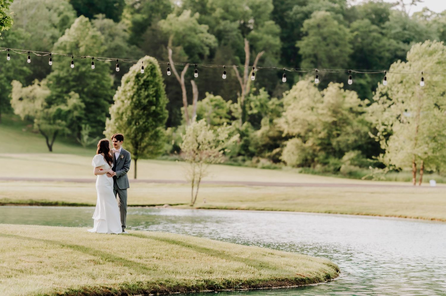 Two figures embrace by a lake on a sunny day with lush green trees in the background at a tranquil park setting.