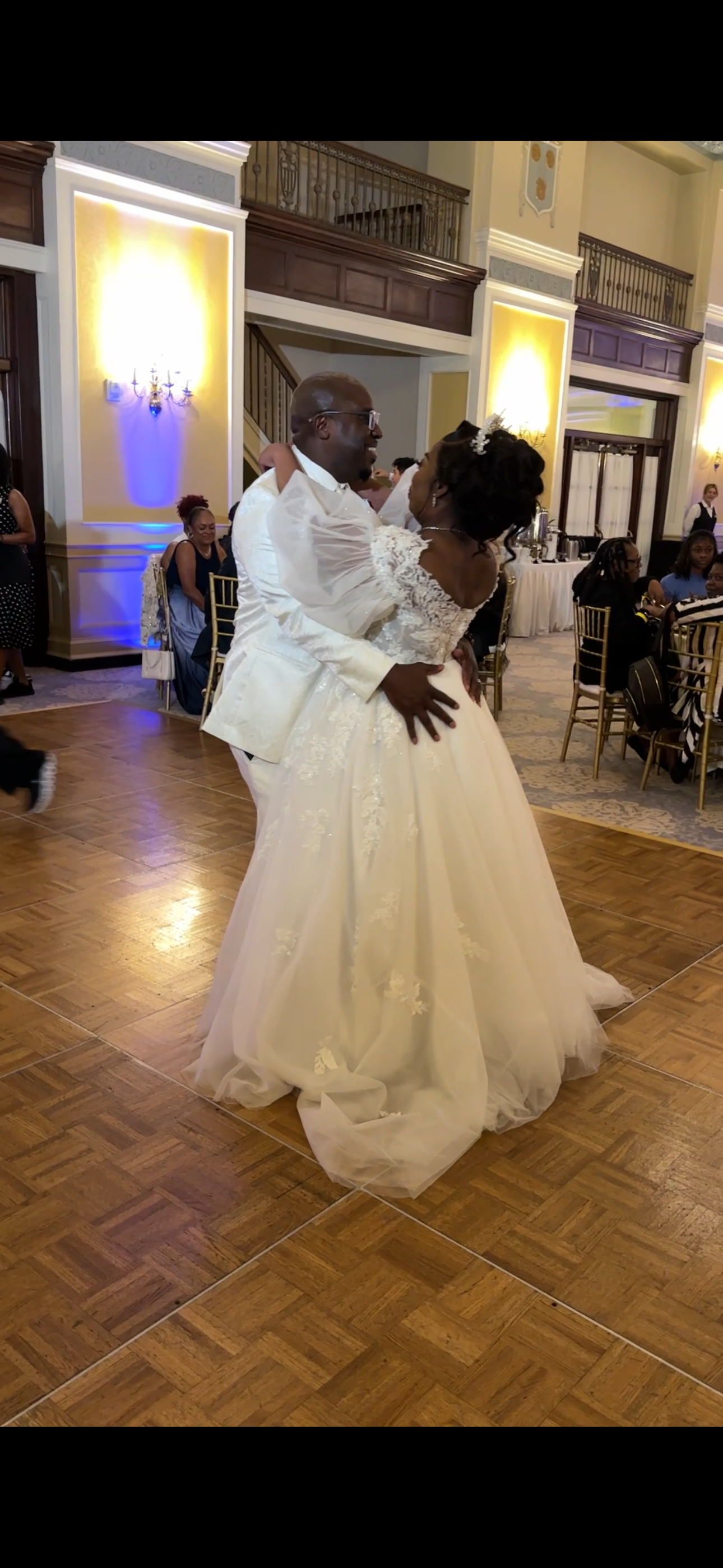 A couple shares their first dance at a wedding reception on a parquet floor in an elegant ballroom with illuminated sconces.