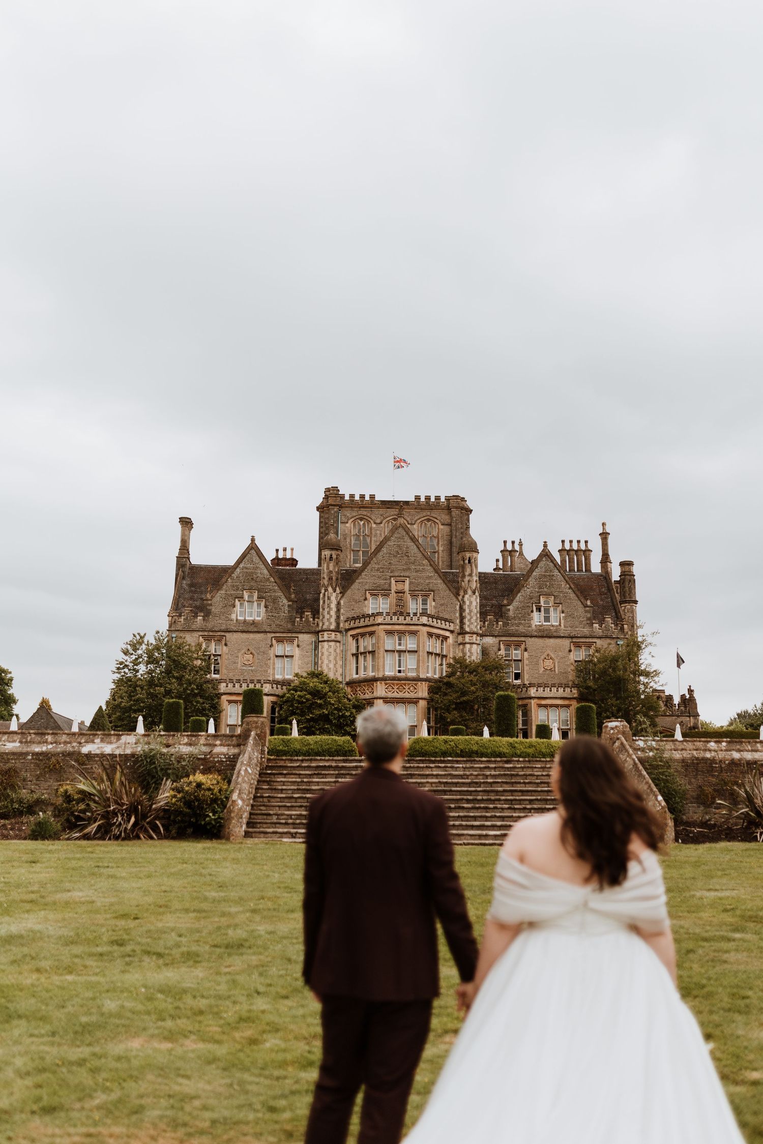 Two people walk up stone steps towards a Tudor-style entrance framed by autumn foliage at an elegant brick building.
