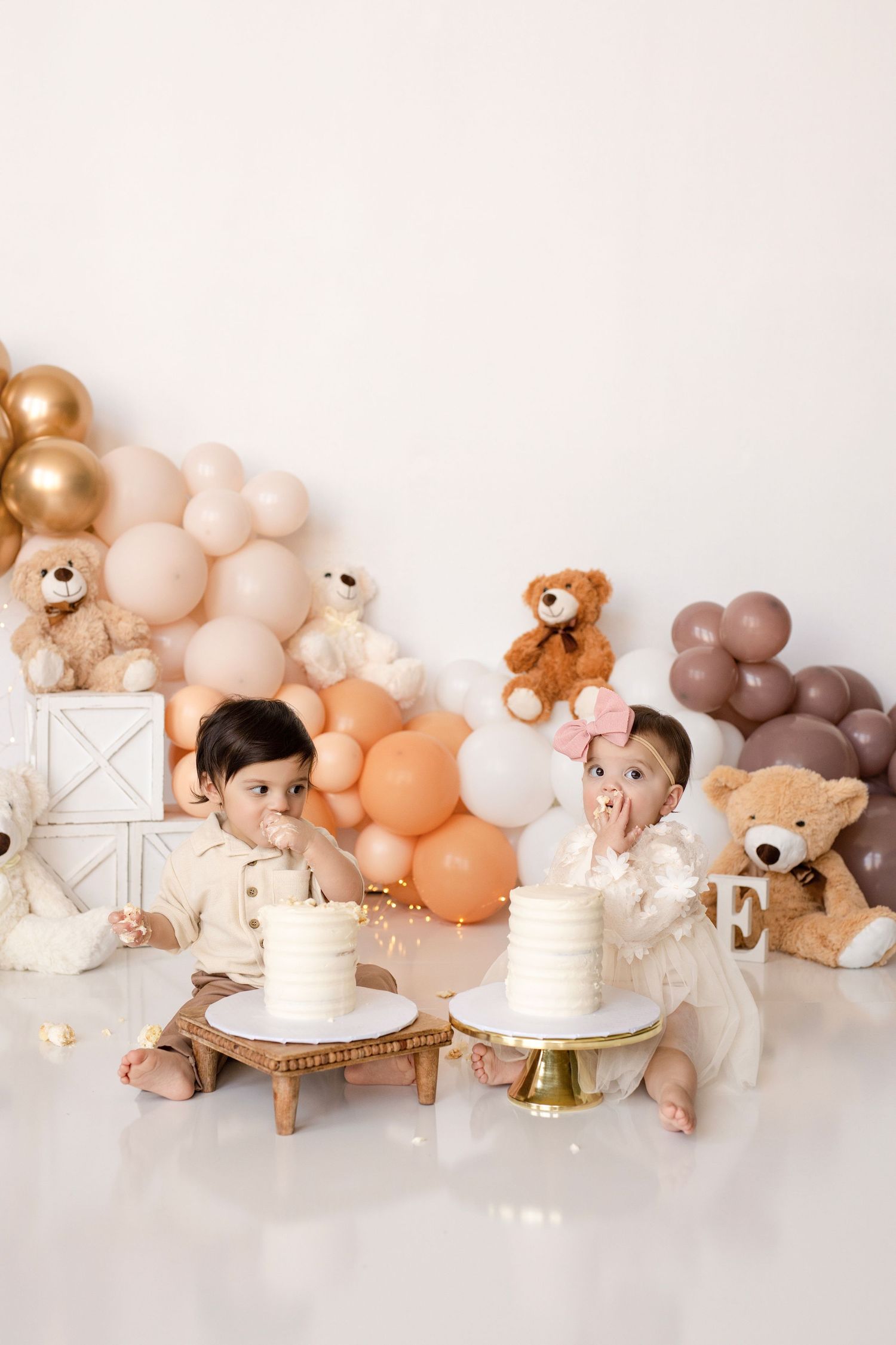Neutral-toned balloon arch with teddy bears and white cakes create a cozy first birthday celebration setup.