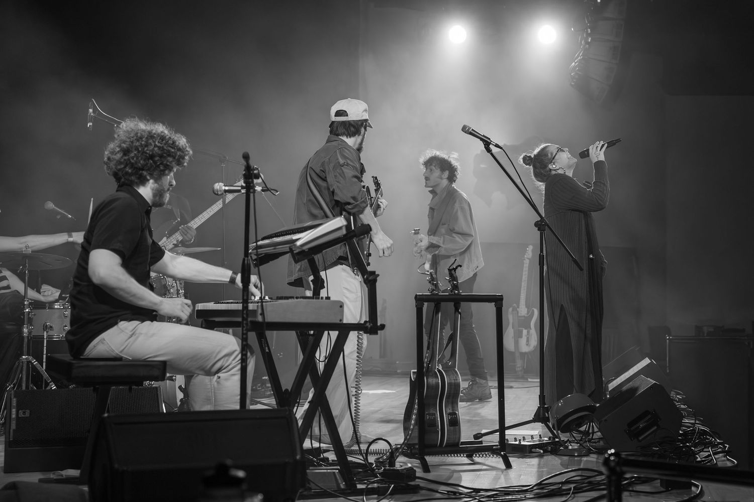 Musicians perform with electronic equipment in atmospheric black and white stage lighting.