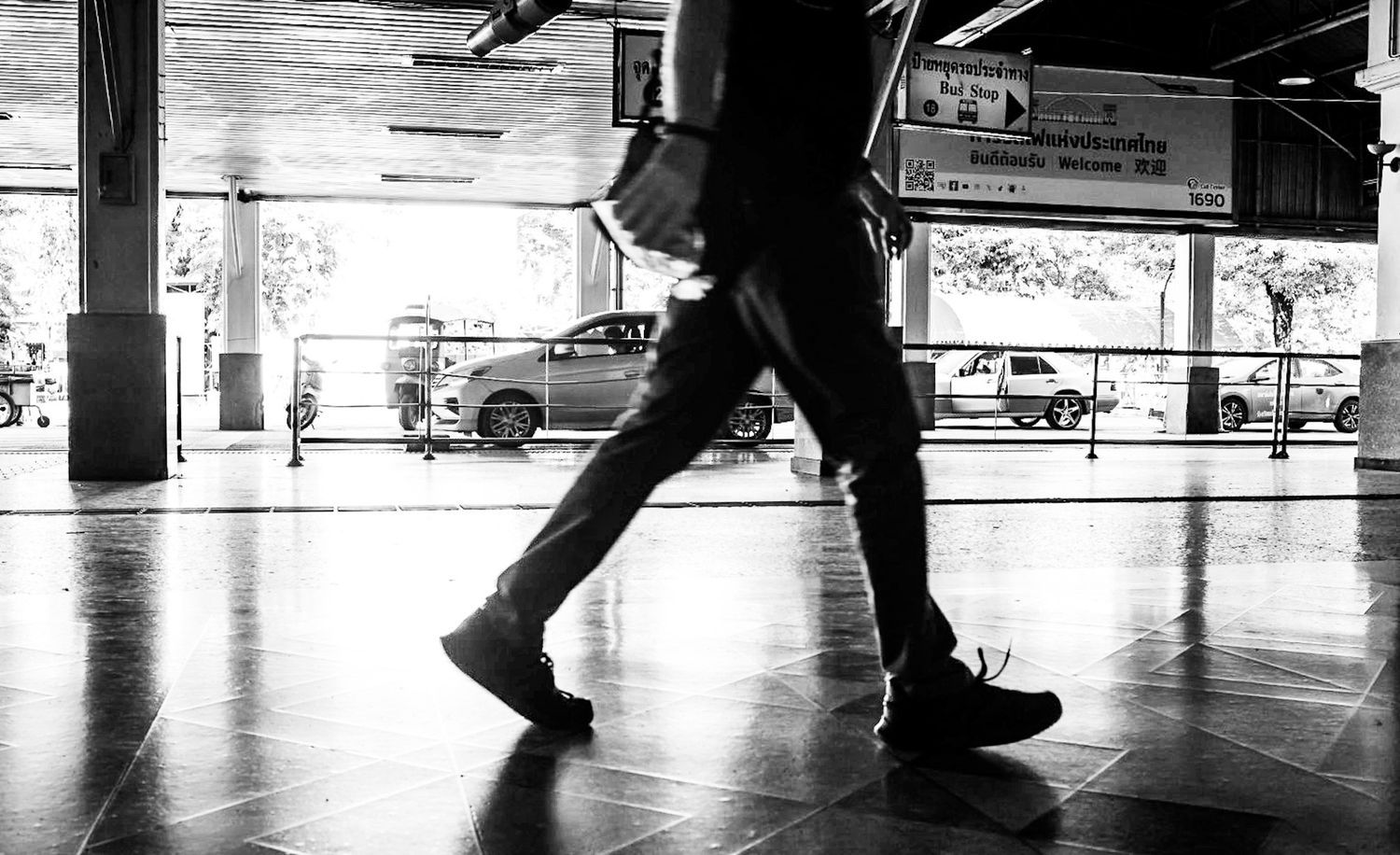 Black and white street photo of a person walking through an open-air covered walkway near parked cars.