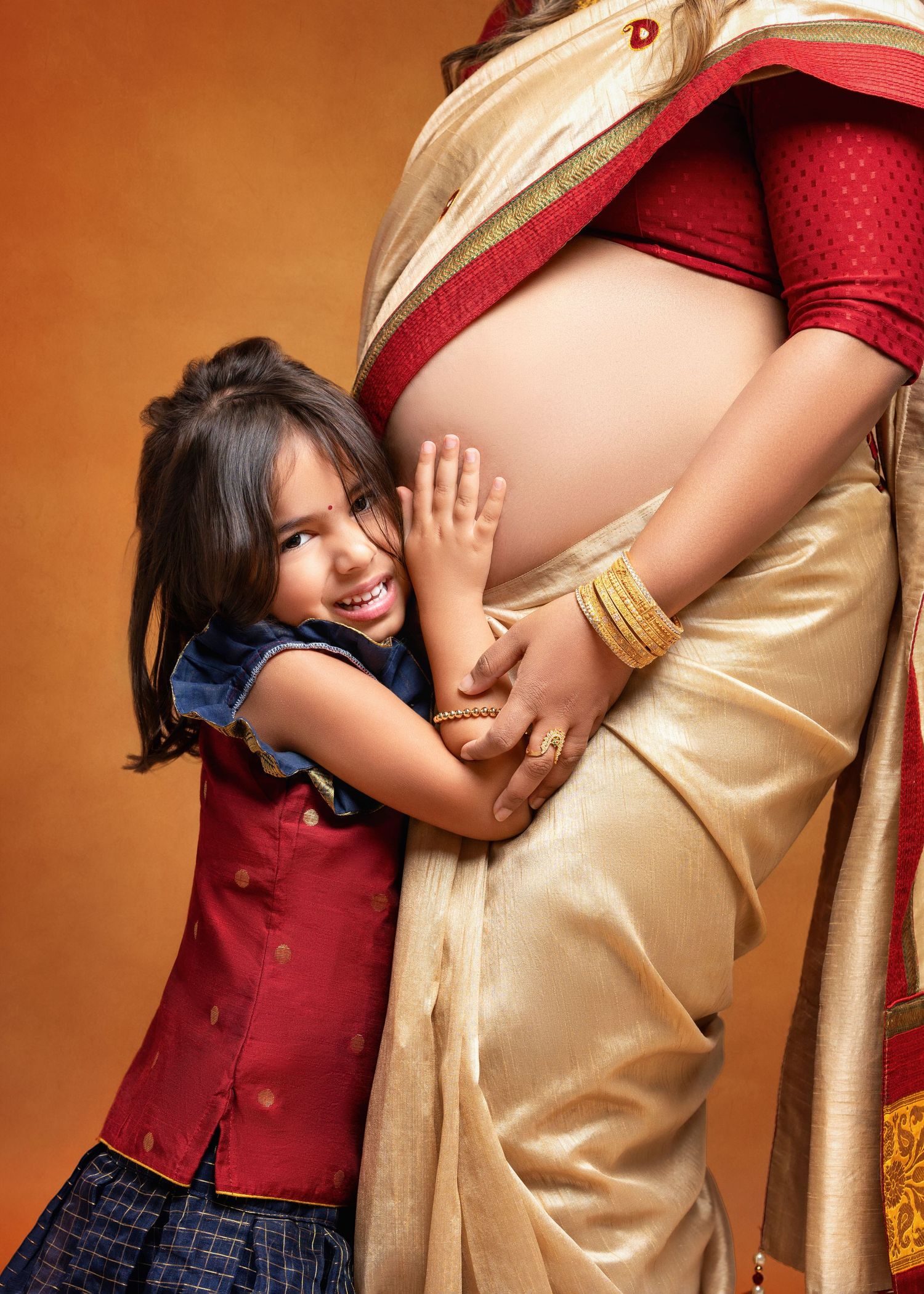 a girl smiles and rests her hand on her mama's belly during their saree maternity photoshoot