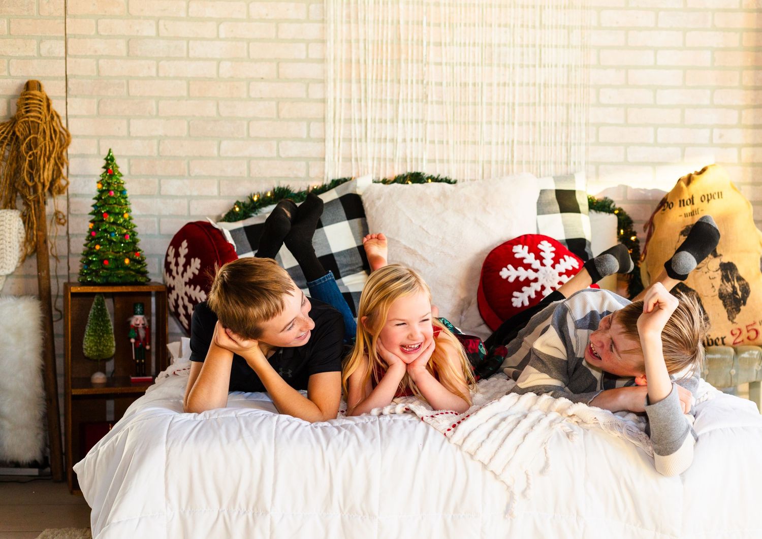 Three people lying on a white bed with holiday decor and buffalo plaid pillows in a festive setting.