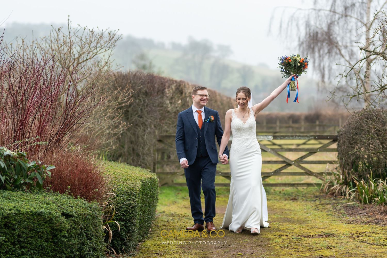 A couple walking through a countryside garden path on their wedding day.